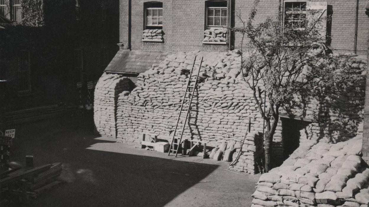 A ladder resting against several sandbags placed in front of a police station.
The sandbags have been stacked to the first window. The image is in black and white.