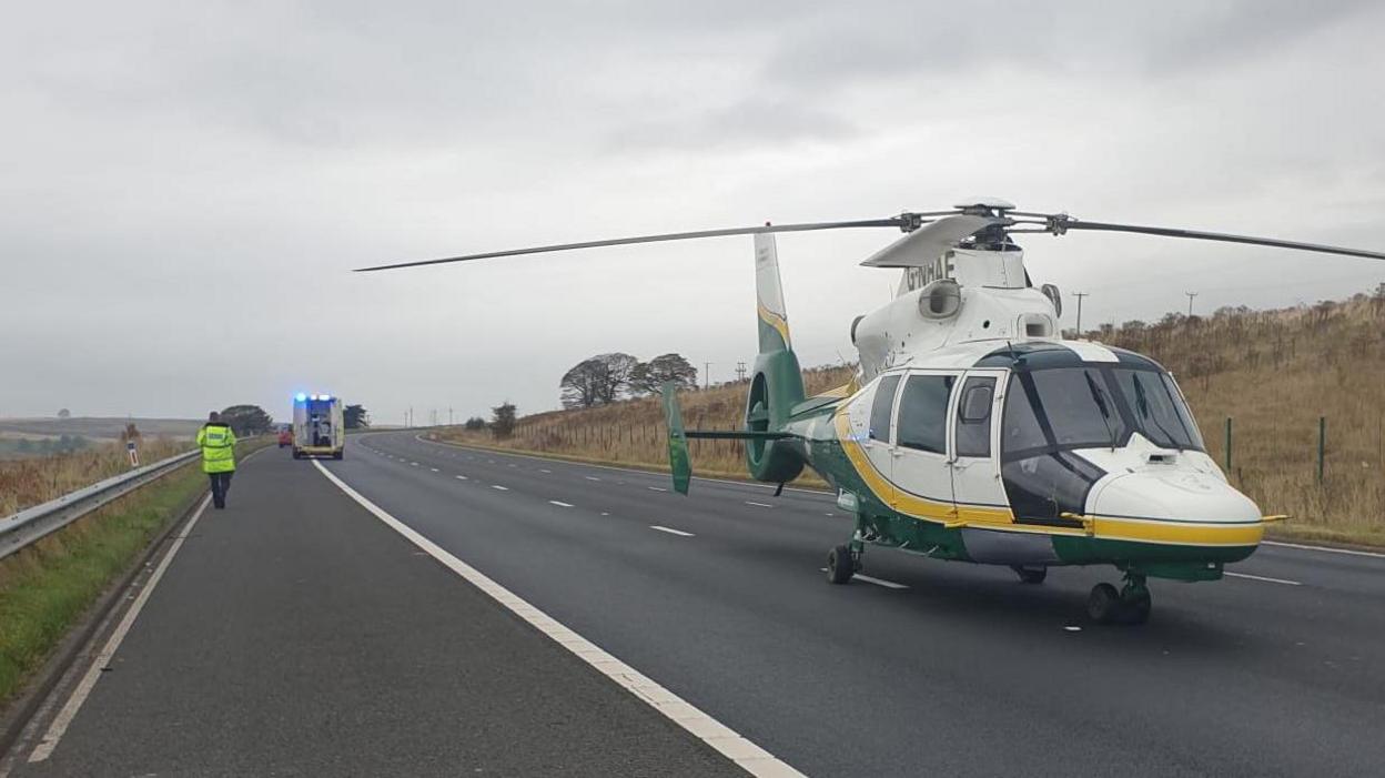 A helicopter parked on a motorway. An ambulance is in the background with a person wearing a hi-vis jacket.