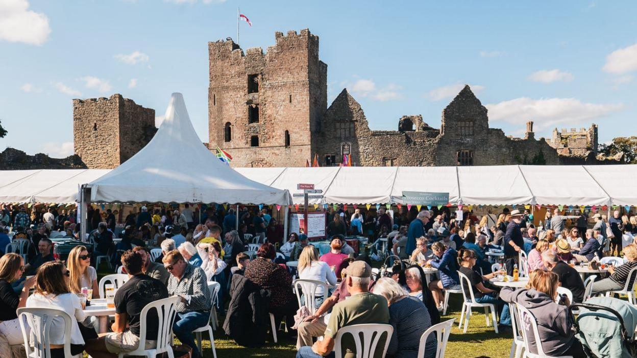 Crowds of people, some of them seated with marquees in the distance and a castle behind them