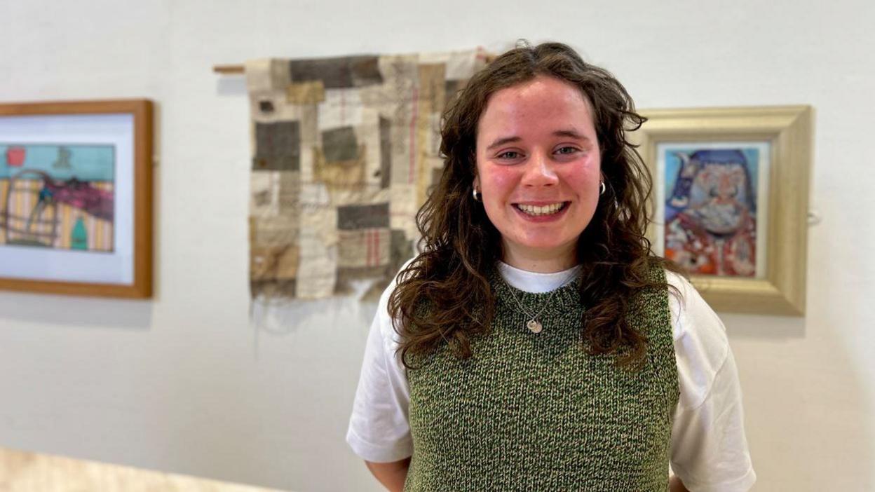 A woman wearing a white and green top stands in front of pieces of art. She is smiling at the camera. Behind her lies two paintings, as well as a piece of patchwork.