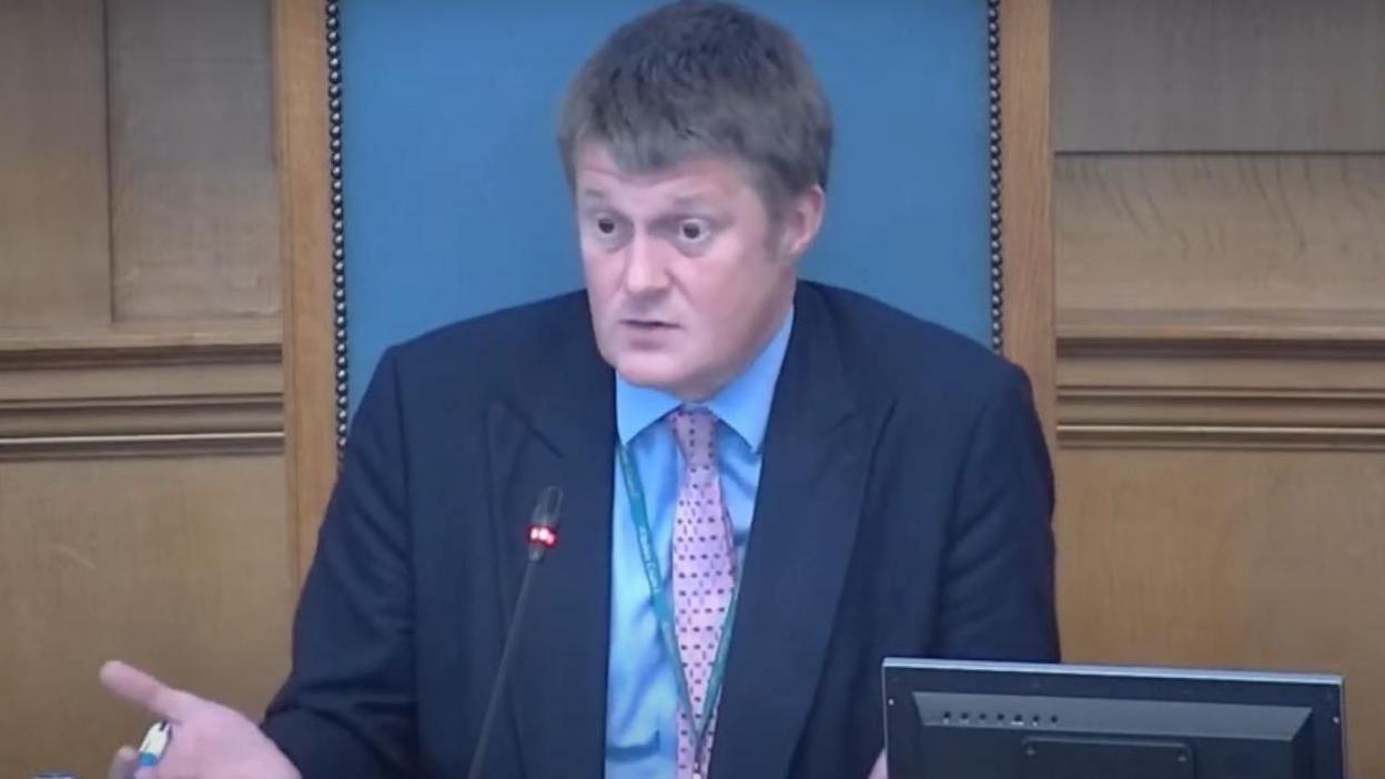 Richard Clewer speaks at a council meeting. He is wearing a navy blue suit jacket with a blue shirt and pink polka dot tie. He sits in front of a computer and a microphone and his right arm is outstretched as he gesticulates. 