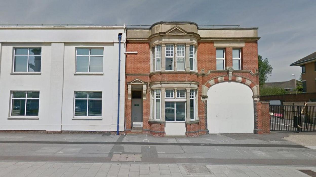 Two buildings on a road. One is red brick and Victorian in style. The other is painted grey.