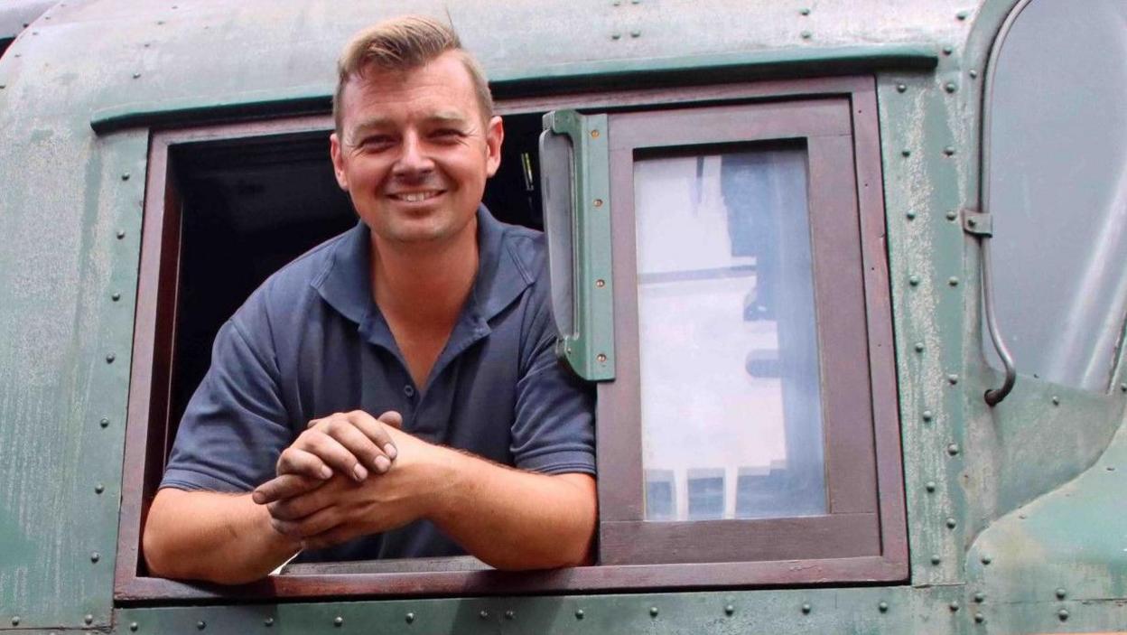 A smiling Billy Johnson leaning out of the window of a locomotive. He has short blond hair and is wearing a blue polo shirt.