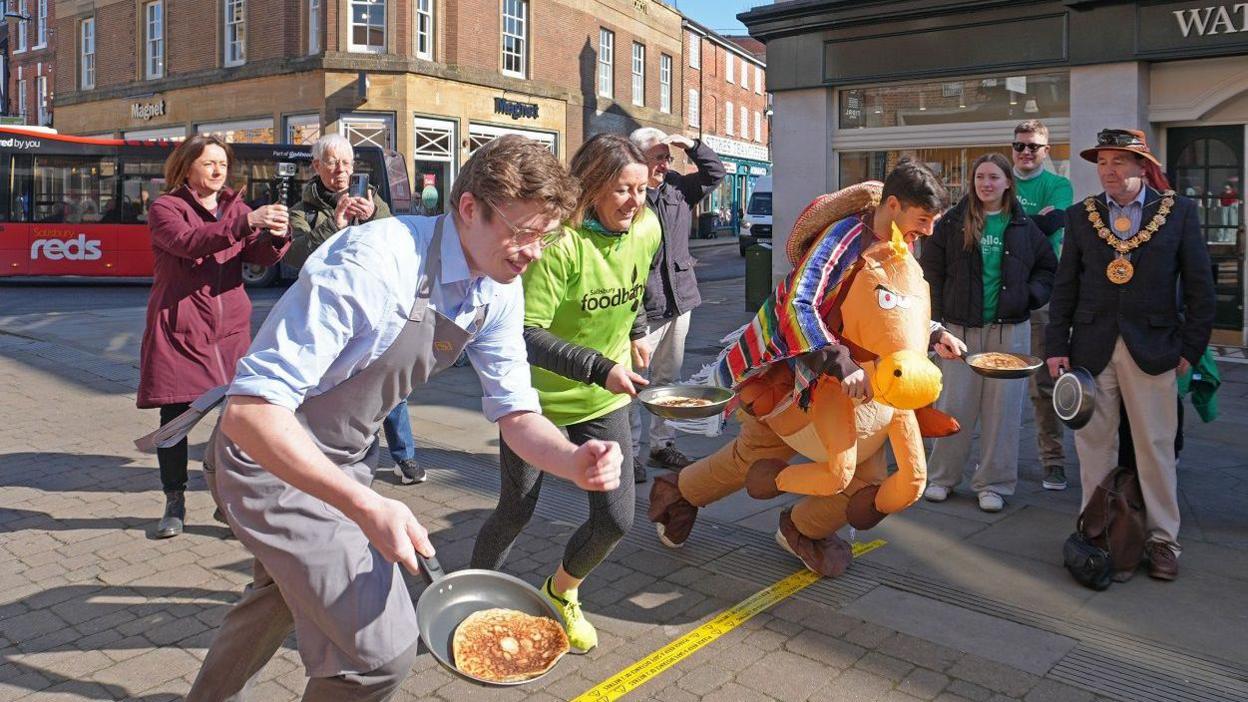 People in a city centre just starting to run over a yellow line taped to the floor in a pancake race. All hold frying pans with pancakes in them. One man is dressed in a horse costume, another man wears an apron. Spectators look on.