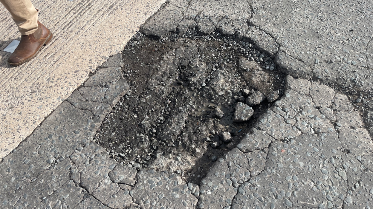 A large pothole in a road. Beside it is a person's leg and foot. They are wearing a brown boot and brown trousers. The pothole is around five times as long as the boot.