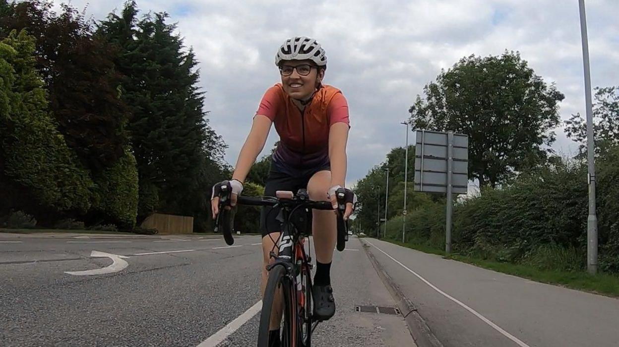 Rebecca Laurel, a 25-year-old woman, riding a bike on a road wearing an orange and purple cycling jersey and a white helmet.