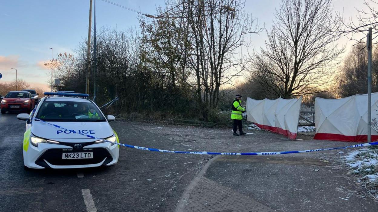 A police car at the scene in Salford, with police tape and a lone police officer standing in front of white screens which appear to be about 7ft (2m) tall.