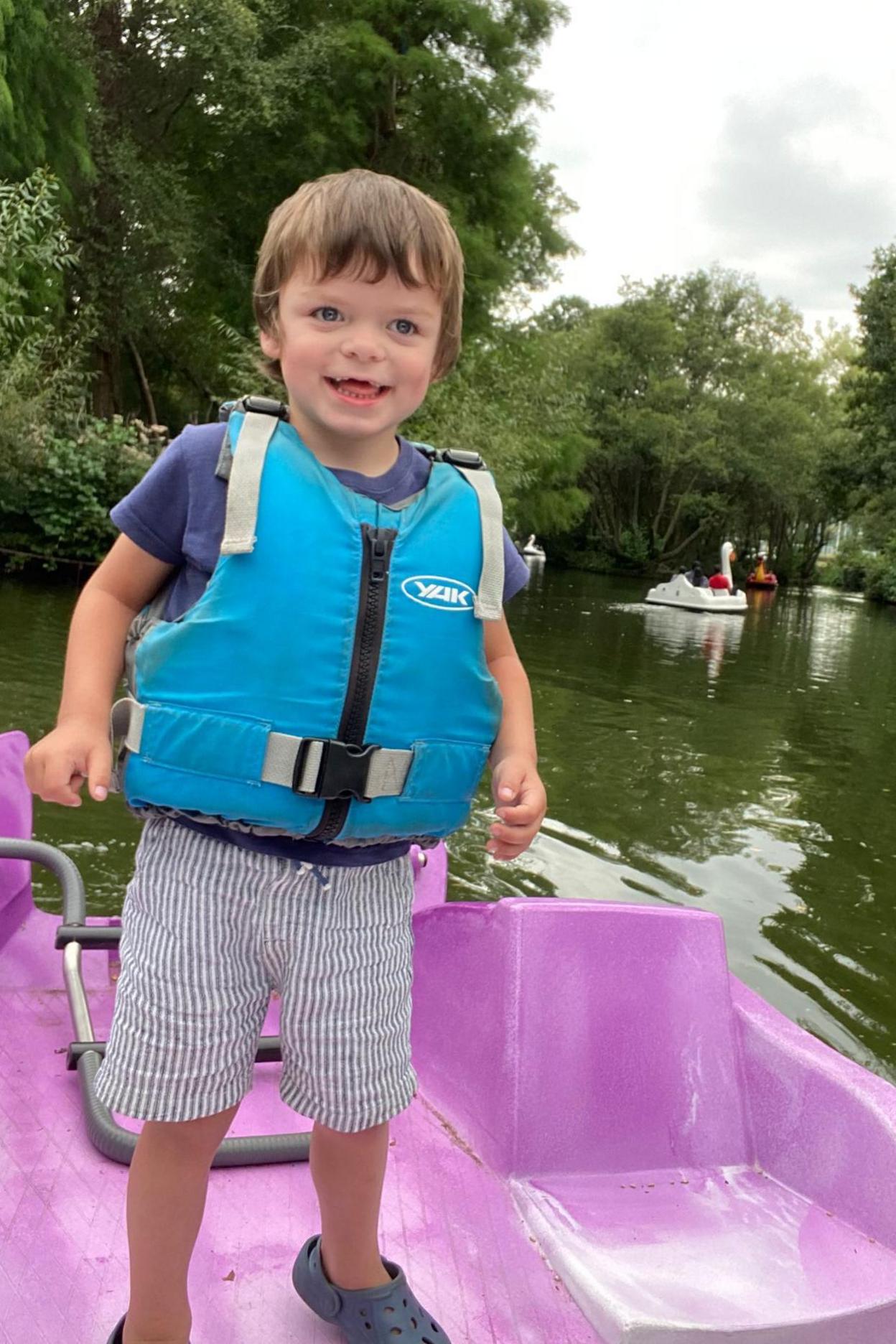 A little boy with light brown short hair is smiling while standing on a pink pedal boat on a lake. He is wearing a blue T-shirt, striped blue shorts and a turquoise life jacket. There are large pedal boats shaped like swans and other birds behind him.