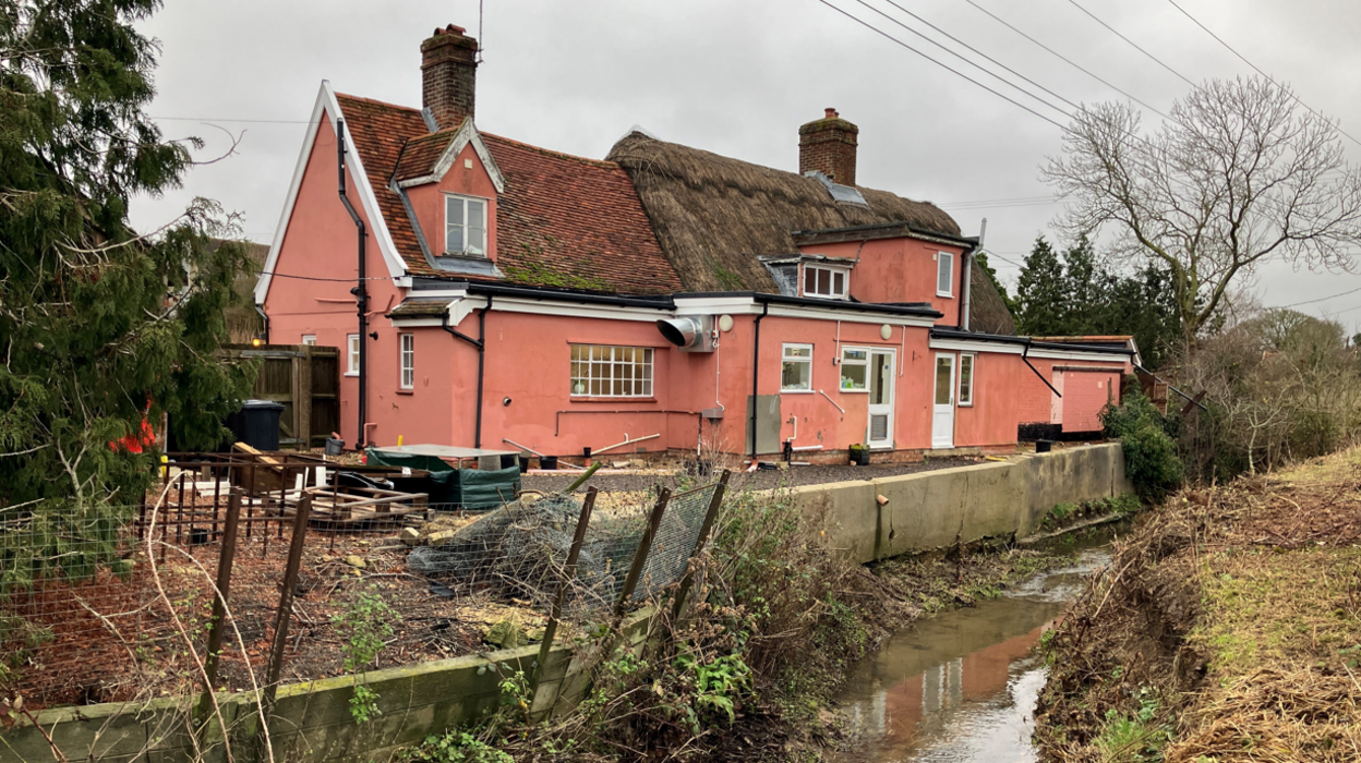 The rear of The Framsden Greyhound - with pink stuccoed walls - backing onto a stream with a concrete retaining wall between the building and the waterway. The area is a little scrubby and messy.