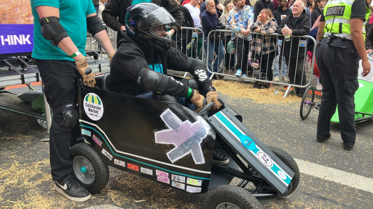 A small black cart is on the road with someone holding it. The person inside of it is wearing a black jumper, elbow pads and a helmet. People are stood behind a barrier watching.