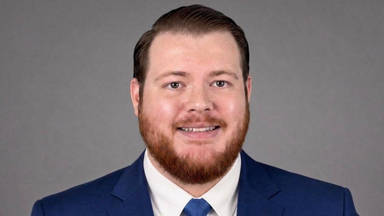A man with short, coiffed hair, and a short ginger beard is smiling at the camera. He is wearing a blue suit and tie. 