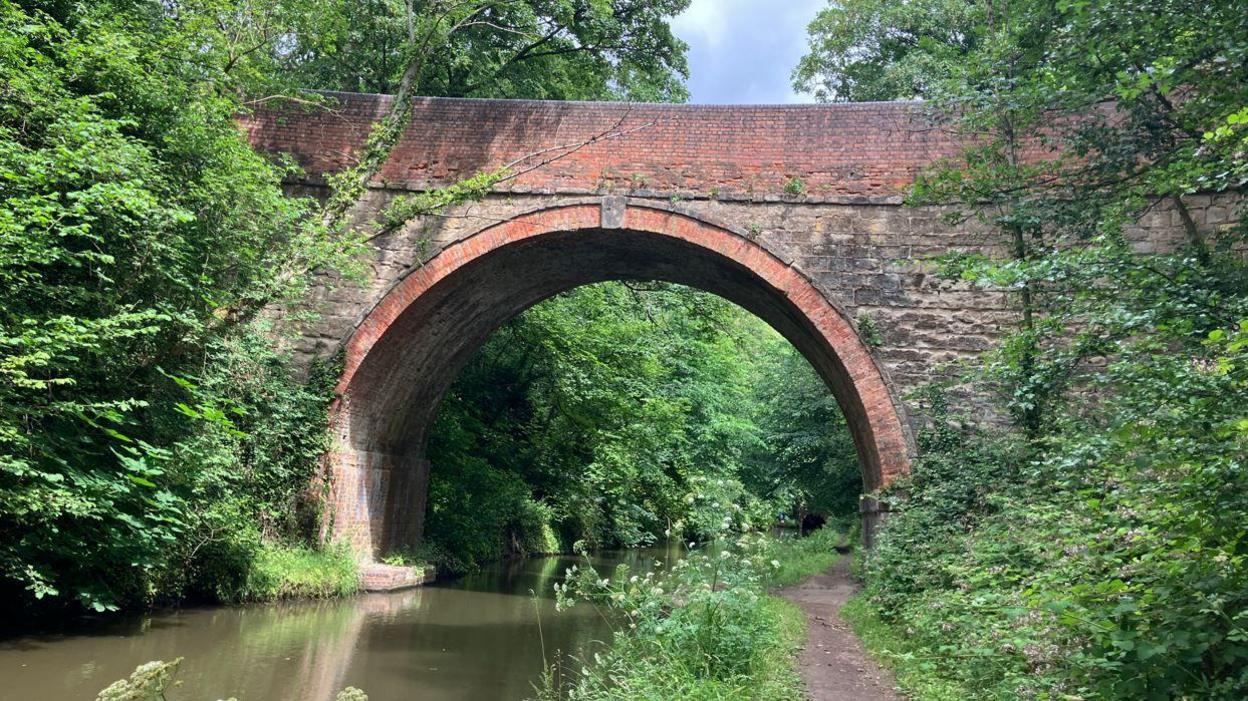 A brick bridge over the canal. A footpath and trees and bushes can be seen on the each side.