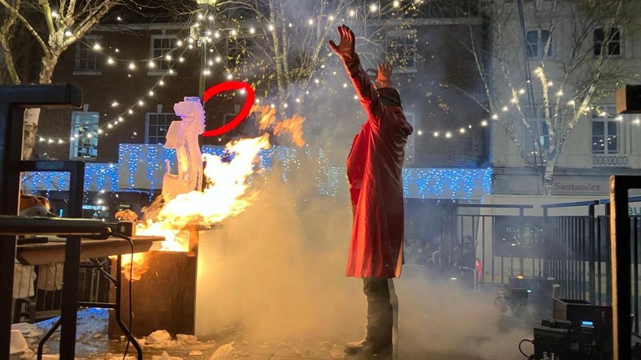 A man in a red jacket with his arms in the air, on stage. He is stood infront of a fire display infront of a crowd watching on