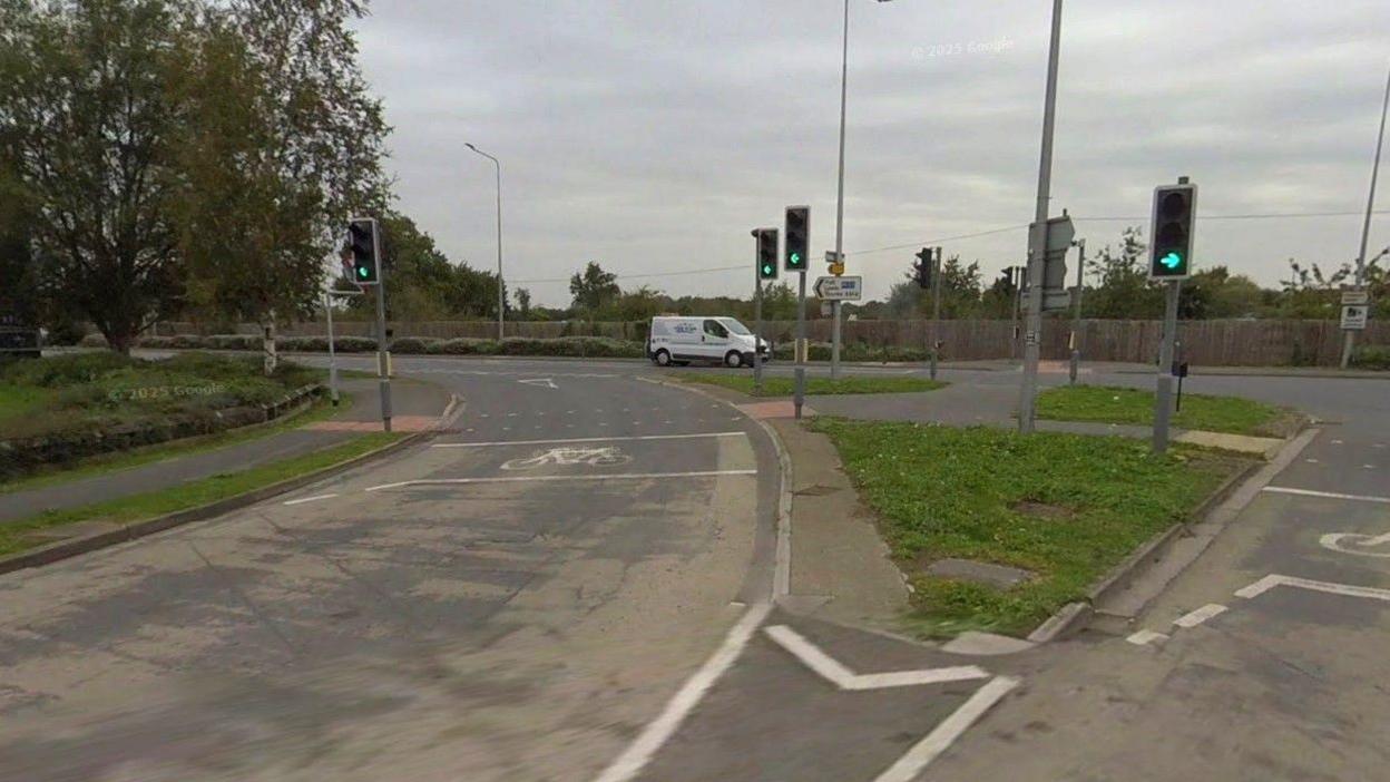 A large road junction with carriageways turning left and right, split by a triangular grass verge and crossing points. Four sets of traffic lights can be seen, all on green. A tree stands to the left. A white van is waiting at lights in the background. The sky is grey.