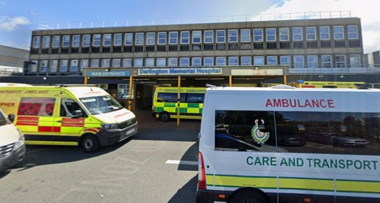 Several green, white and yellow ambulances parked outside Darlington hospital. It it s a large flat grey building with two longs rows of windows above a canopy over a front door.