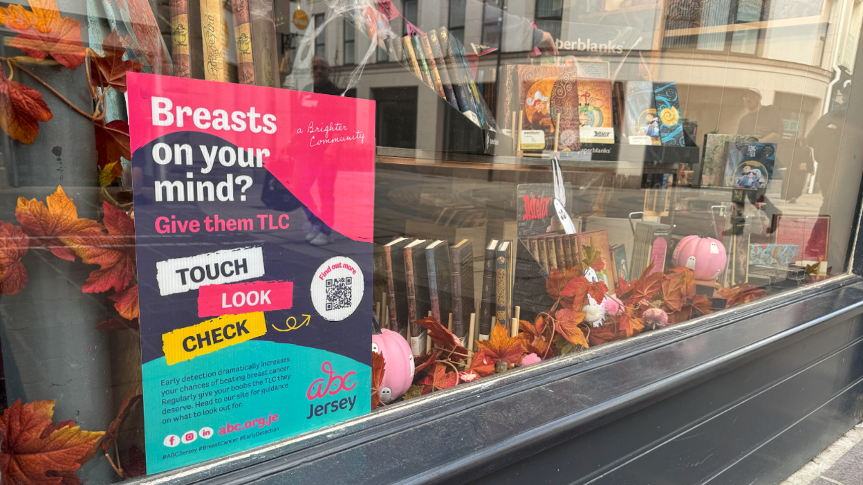 The photo shows a shop window with an ABC Jersey poster explaining how to check your breasts. The window display has pink painted ghost pumpkins and surrounded by brown autumn leaves around its display of books.