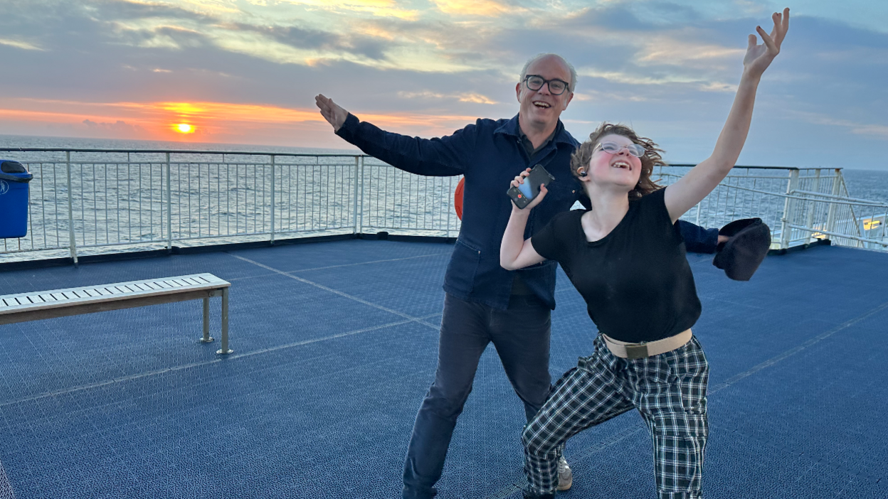 Julian Francis and April, smiling and throwing their arms in their air on board a ferry at sunset. Mr Francis wears black-rimmed glasses, a navy shirt and jeans. April wears glasses, a black t-shirt and chequered trousers, holding a phone in one hand.