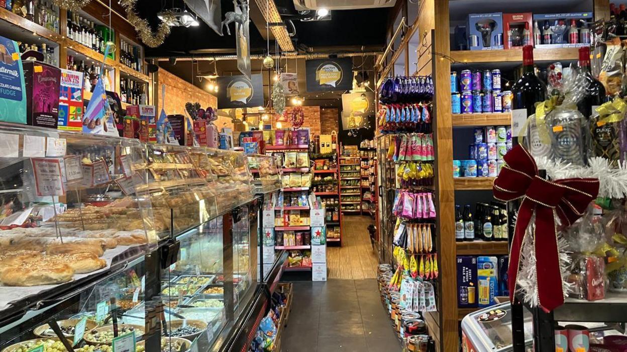 A delicatessen counter on the left hand side of a heavily stocked independent grocery store filled with seasonal products, food and drink