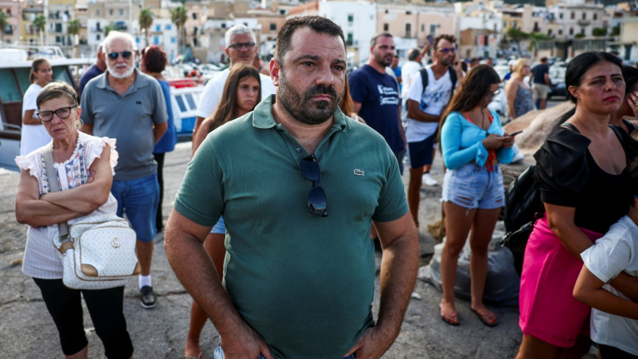 People gather in the coast town of Porticello as rescue personnel transport a body bag after a luxury yacht