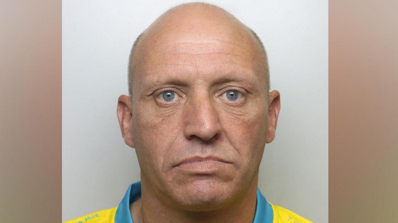 A bald man in his 40s, wearing a yellow top with a blue collar which looks like a football shirt. He is staring into the camera.