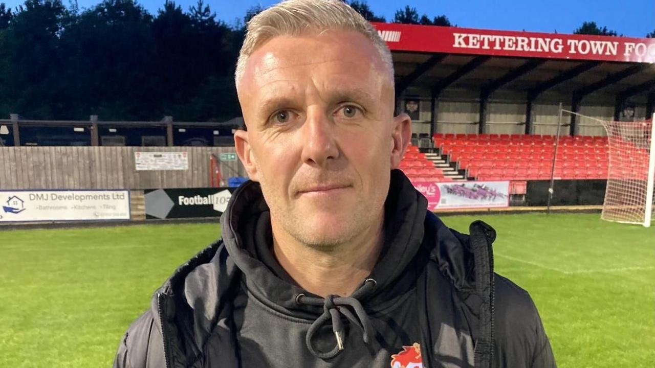 Richard Lavery, who is wearing a black hoodie and black coat. He has short grey hair and is standing in front of a stand at the Kettering ground, which has red seats.