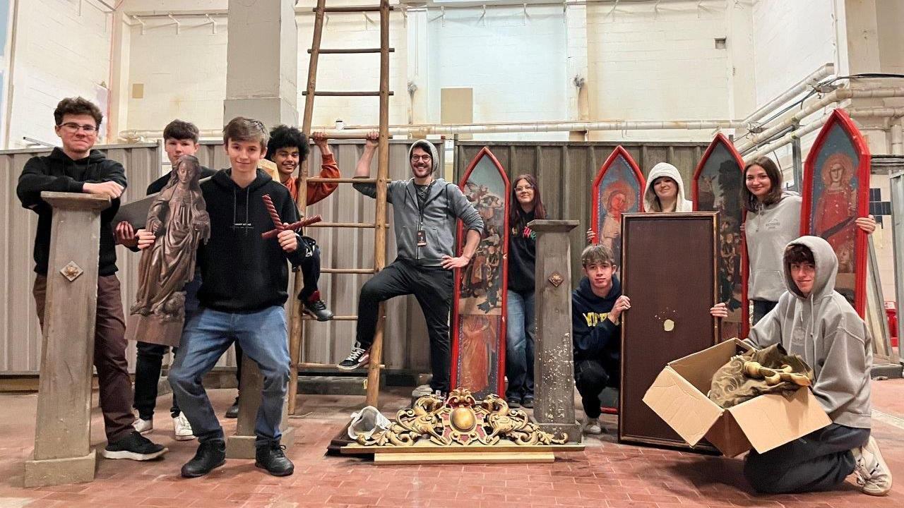 A group of nine film students posing with items from the sets of the BBC drama Wolf Wall including a plynth, paintings and a ladder.