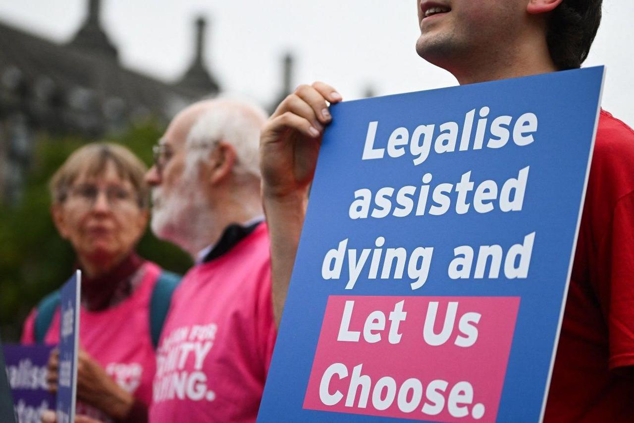 A campaigner from "Dignity in Dying" holds a placard during a demonstration outside The Palace of Westminster. The sign is blue with write writing and says: "Legalise assisted dying and Let Us Choose".