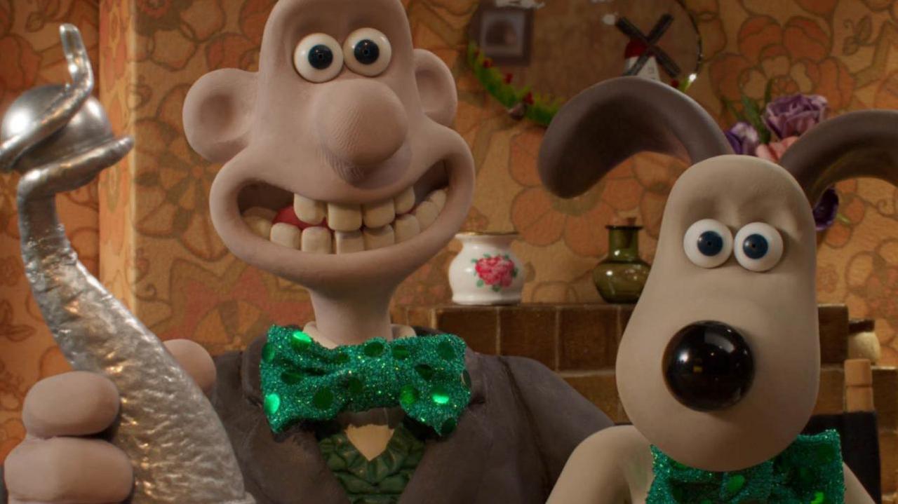 Image of clay modelled characters Wallace and his dog Gromit.  Wallace is holding a silver trophy.  They are both wearing green shiny green bow ties.  In the background can be seen a tile fireplace, oval mirror above the fire and a china and glass vase.
