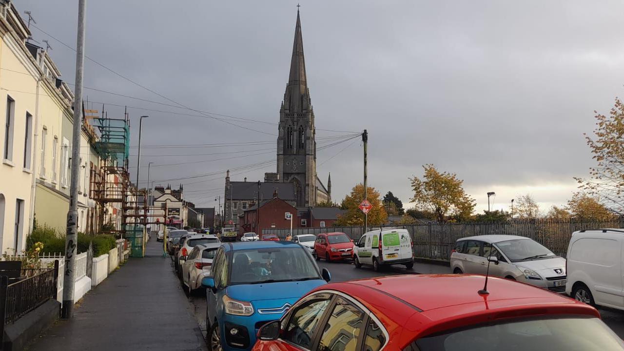 A row of terraced townhouses with a long line of parked cars outside each of them. There is a long line of cars parked on the other side of the street also. A cathedral can be seen in the background