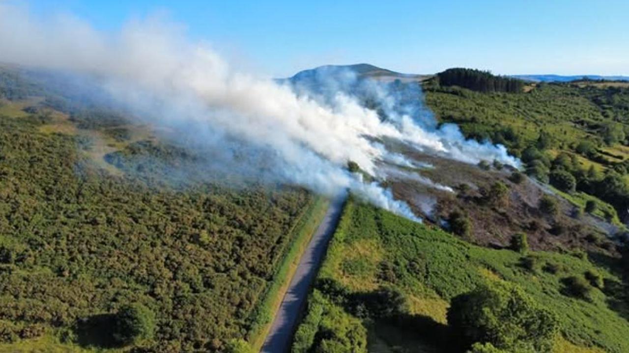 An aerial view of hills, with various pockets of smoldering bush sending white smoke high into the blue sky