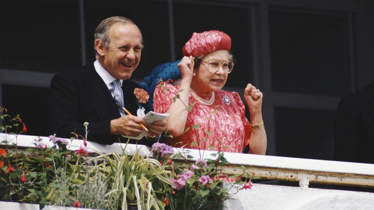Queen Elizabeth II with looking excited with her arms up at the derby races, next to her there's Sir William Heseltine holding a notepad in his hands.