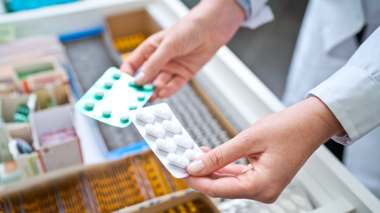 Hands holding two blisters of tablets over a counter with a range of products in it