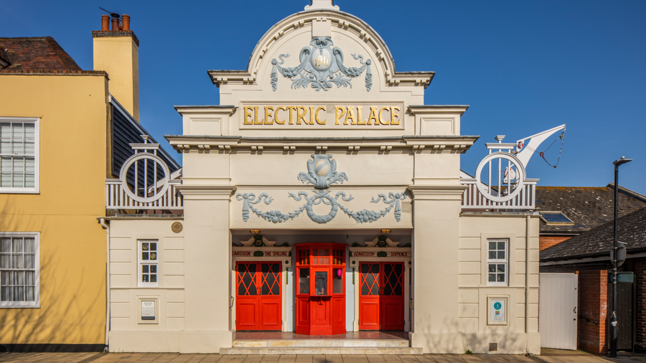 Edwardian cream-coloured building with red doors and Electric Palace in bas relief gold lettering above the door. The building is decorated with plasterwork of wreaths and vines, painted grey.