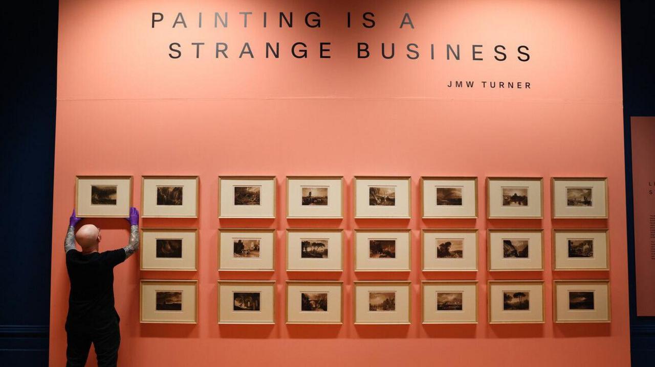 A staff member places a painting on a peach coloured wall covered with paintings.