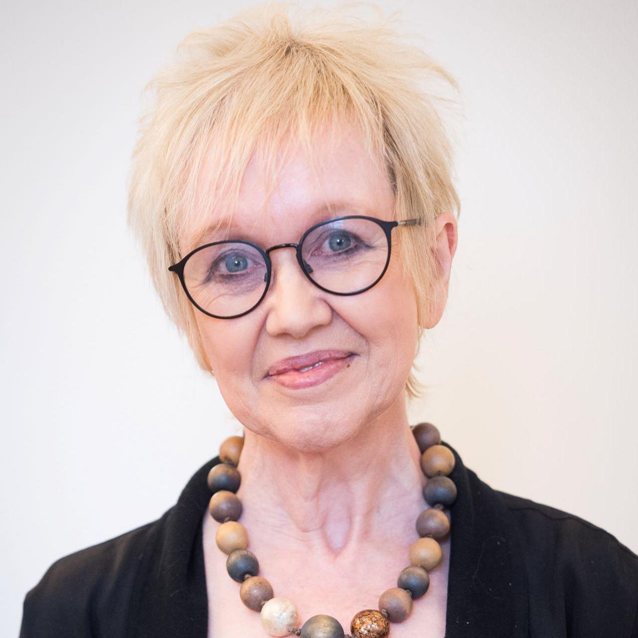 Dr Jane Morris looking straight at the camera in front of a plain white background. She is wearing black framed glasses, a black jacket and brown wooden beads.