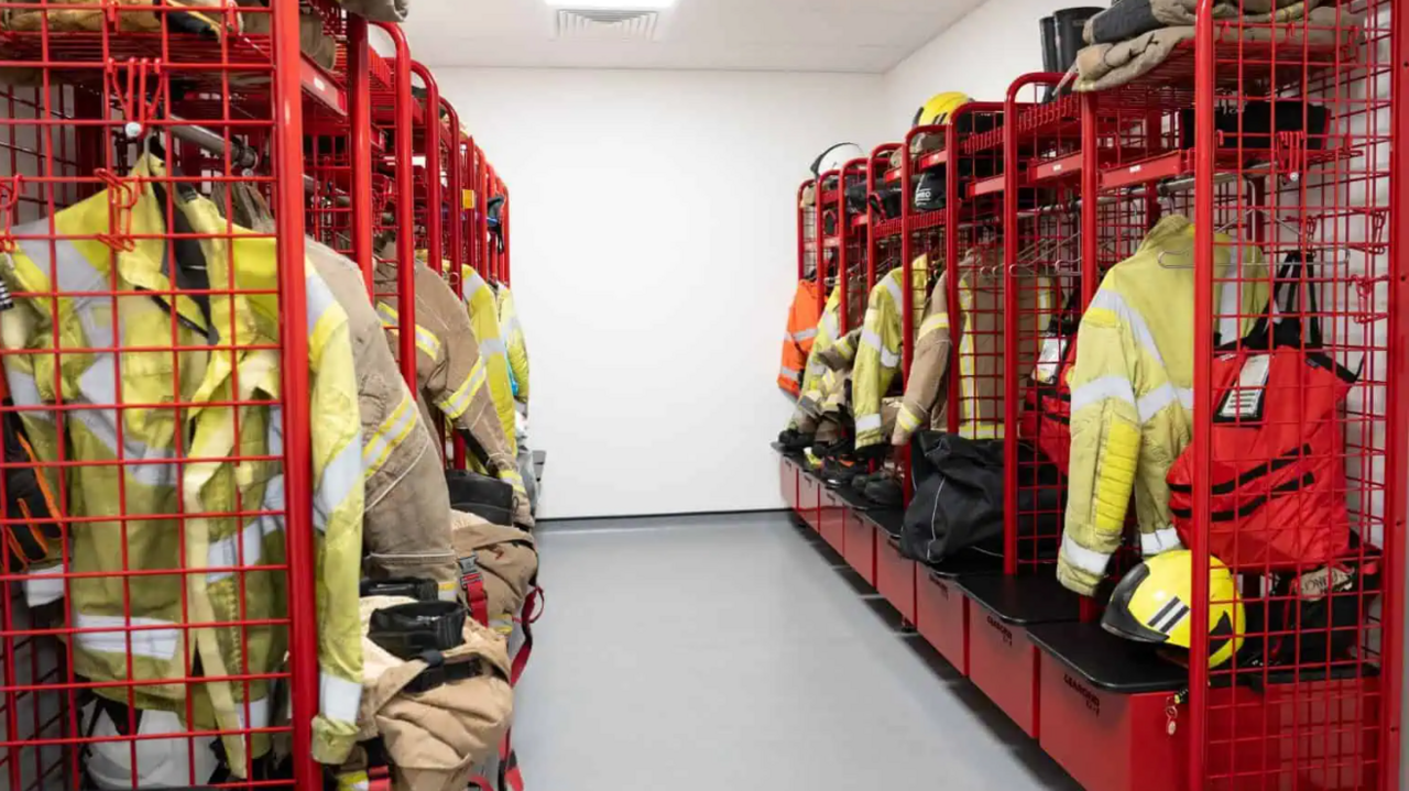 A windowless room with white floors, walls and ceilings, with high-visibility jackets, helmets and other firefighter clothing hung up on red shelving units on either side.