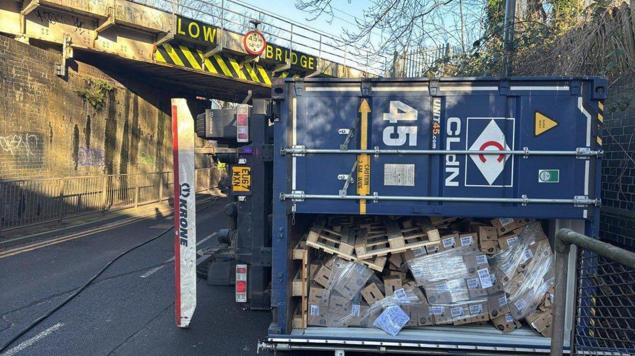 A blue lorry lies on its side on a road underneath a bridge labelled "LOW BRIDGE". Its rear door has opened and pallets and boxes can be seen inside.