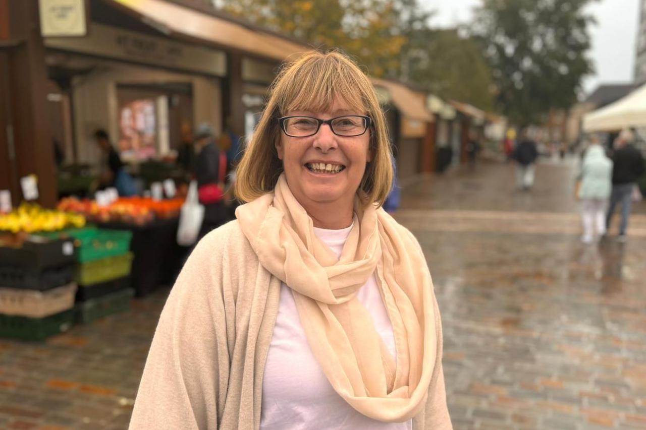 Councillor Wendy Randall standing in Northampton Market Square. She is wearing a cream coloured cardigan and scarf. The market stalls can be seen behind her.