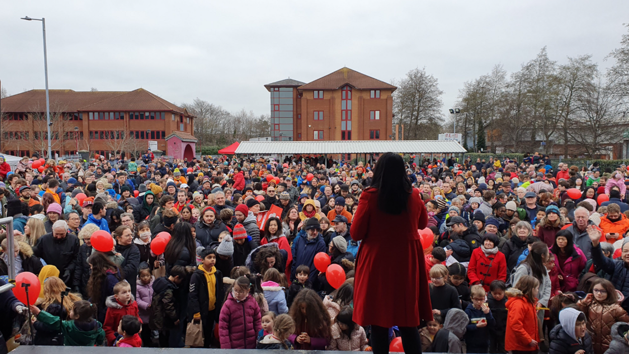 A crowd of people on a winter's day gathered together in front of a stage on which there is a woman in a red coat with her back to the camera