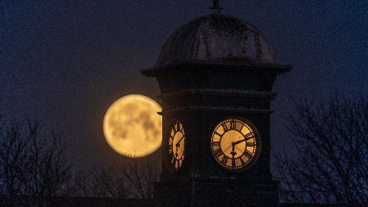 A bright yellow full Moon next to the round face of a clock tower showing the time at 6.10