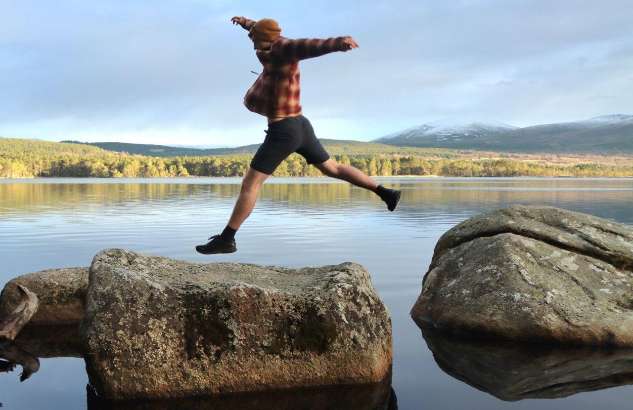 A man wearing shorts, a checked shirt and a hat jumps over rocks with a loch in the background.