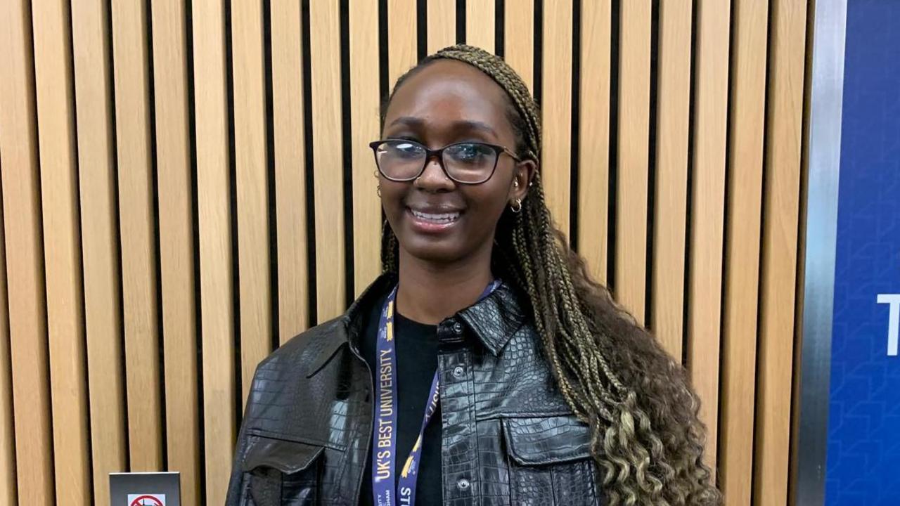 Siyon is smiling at the camera. She is wearing a black t-shirt with a black crocodile leather jacket and small silver hoop earrings. There is a blue and yellow university lanyard around her neck. She has black glasses and long blonde braids. Behind her is wood panelling.