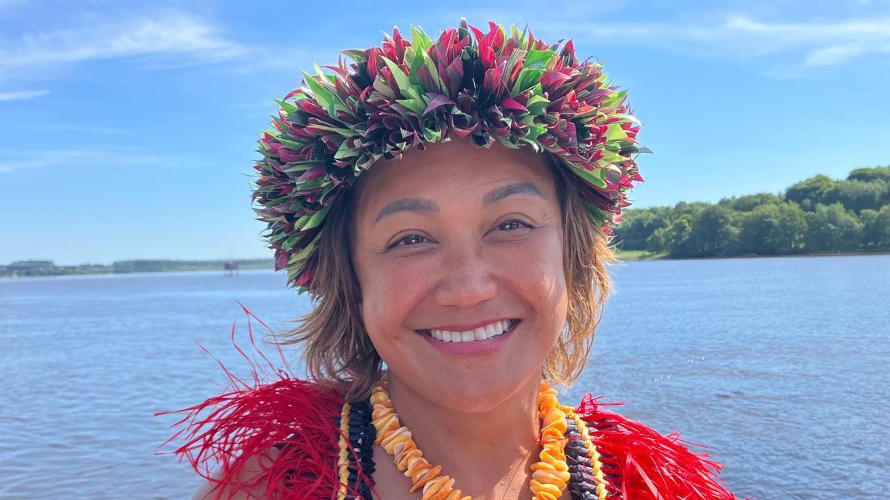 A smiling Dr Melissa Taitano, in traditional brightly coloured head dress and necklaces, stands on the shores of the River Foyle