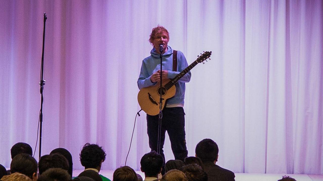 Ed Sheering on stage in front of school pupils. he is holding a guitar and is wearing a lights blue hoodie and dark trousers