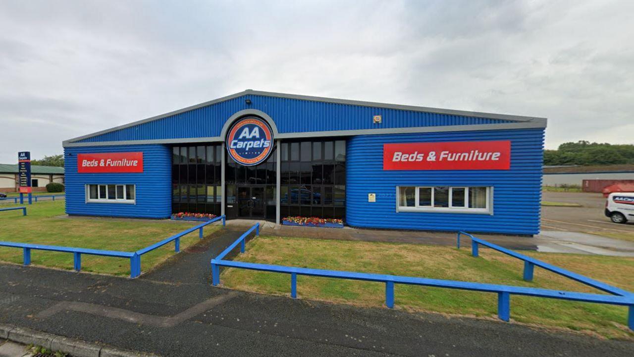 AA Carpets. The building is painted blue with red signs stating it sells beds and furiture. A circular sign also shows the company's name. There is a grass lawn outside the building which is surrounded by a small, wooden fence which has been painted blue.