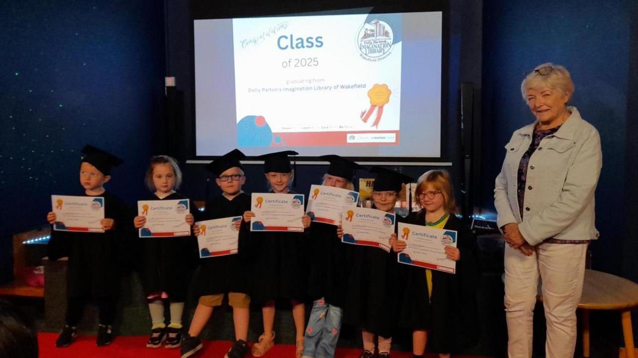 Seven young children stand in a row holding certificates, and they wear graduation gowns and hats, with a screen in the background saying 'class of 2025' and a woman standing to one side, smiling 