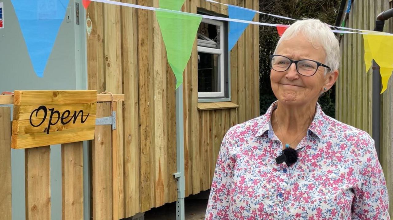 Judy stood in front on the shop. She has short grey hair, glasses and is wearing a pink and white floral shirt.