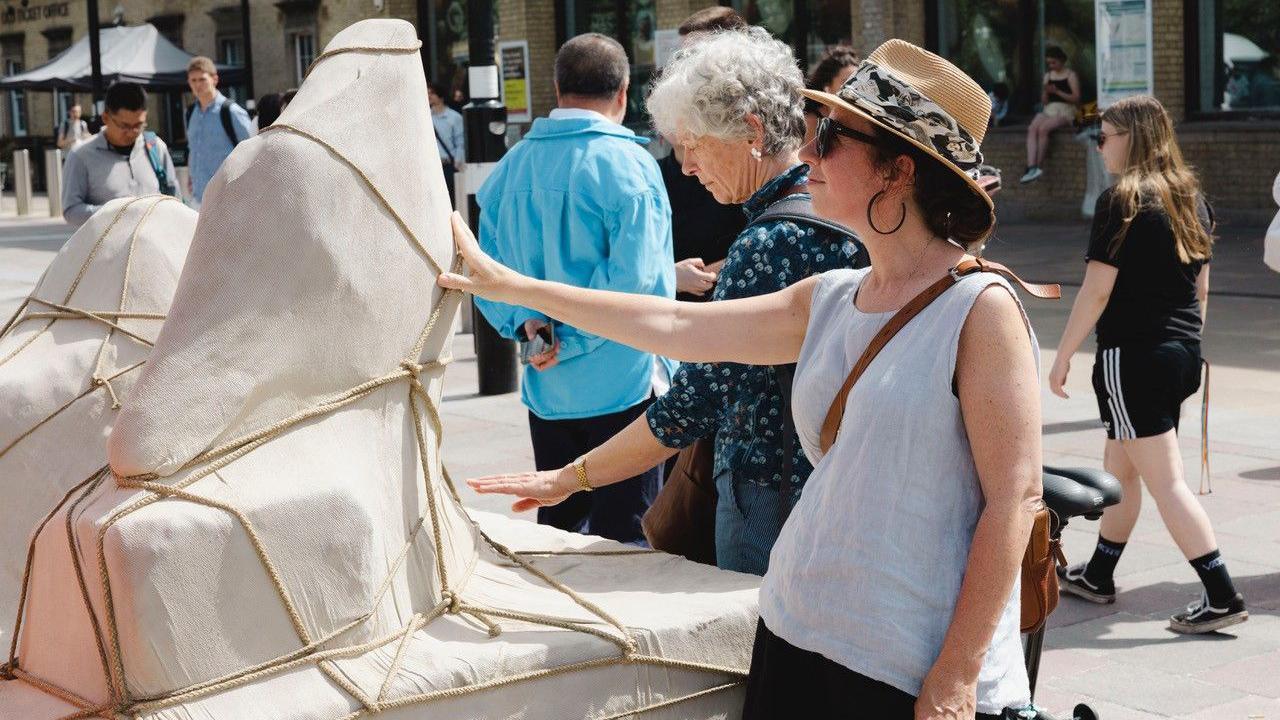 A woman wearing a hat and grey sleeveless top touches the Ariadne sculpture, which is made of grey stone and covered in rope. Other people are looking at the sculpture in the street scene.