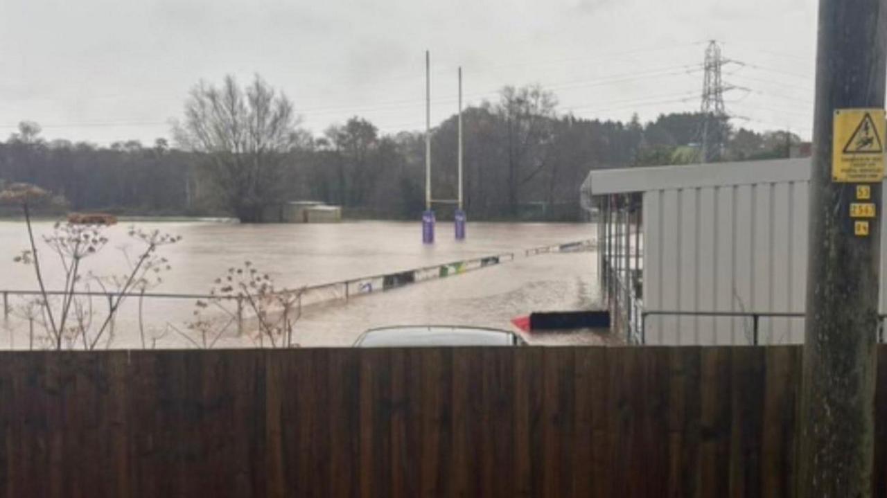 A rugby pitch under a few feet of dirty water. You can see rugby posts in the distance and the top of metal railings around the pitch. There's a brown fence in the foreground there are trees beyond the pitch.