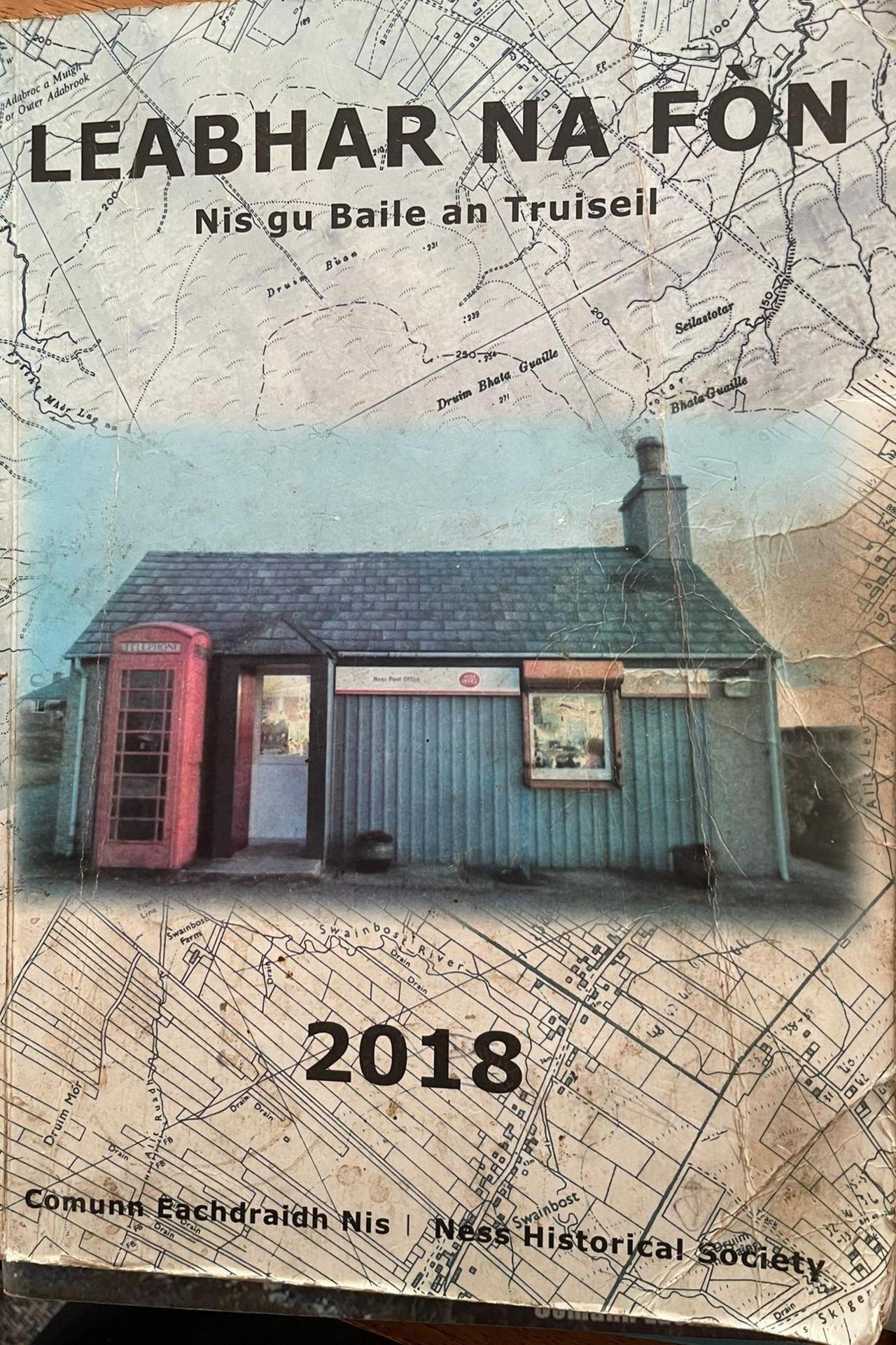 A small, single-storey post office and a red phone box are pictured on the front cover. Lights are on inside the post office. The cover is slightly creased. There are words in Gaelic and the date 2018.